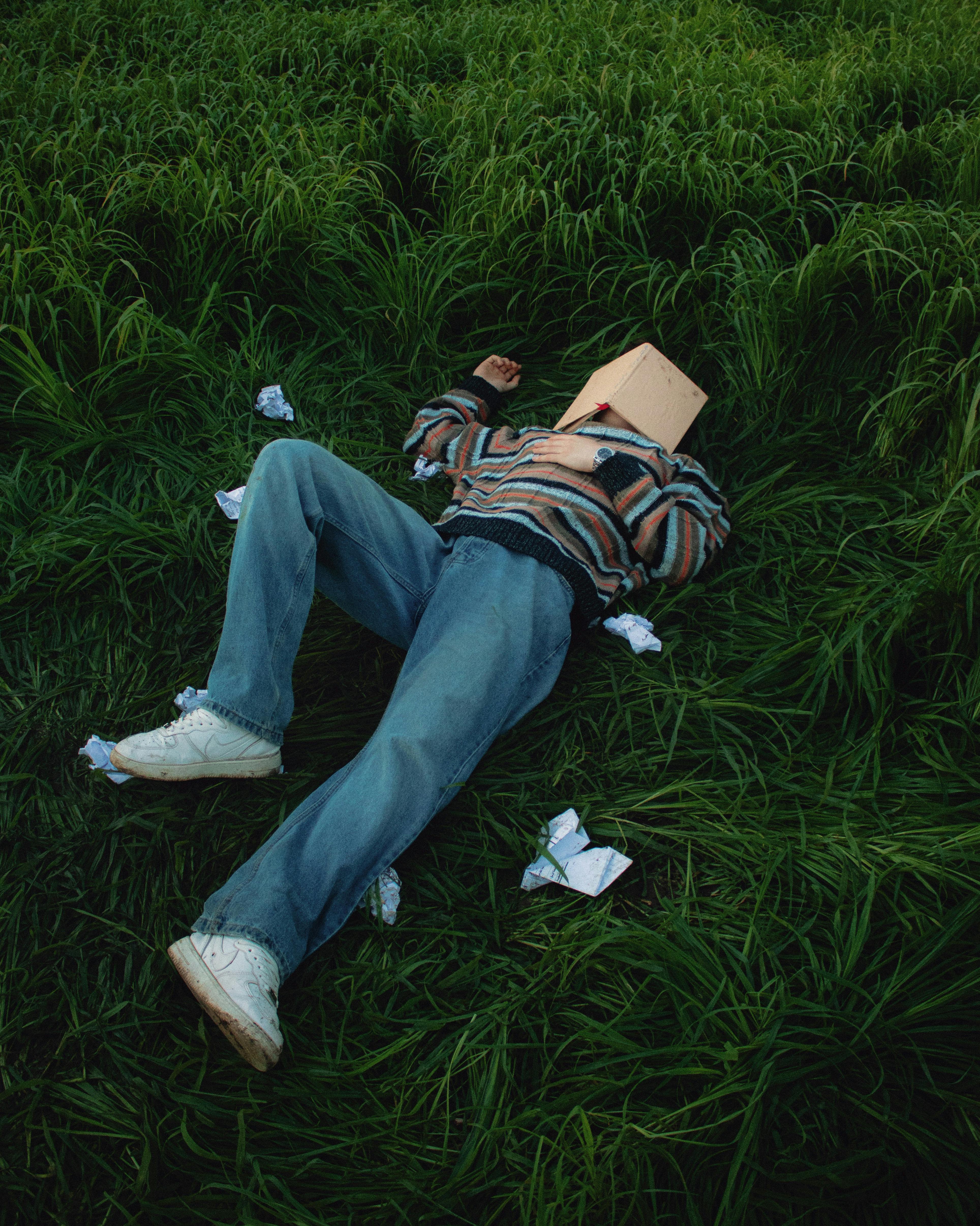 Person lying in tall grass wearing a striped sweater and book over their head, surrounded by crumpled papers